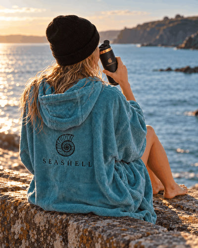 Swimmer in a teal Seashell towelling robe drinking from the Black Flip Top Flask on a harbour wall